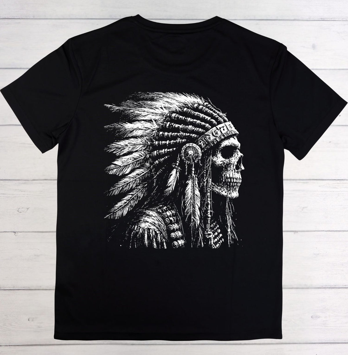 A black short-sleeve t-shirt laid flat on a white wood plank background, featuring a large white graphic print of a skull wearing a feathered headdress on the front.