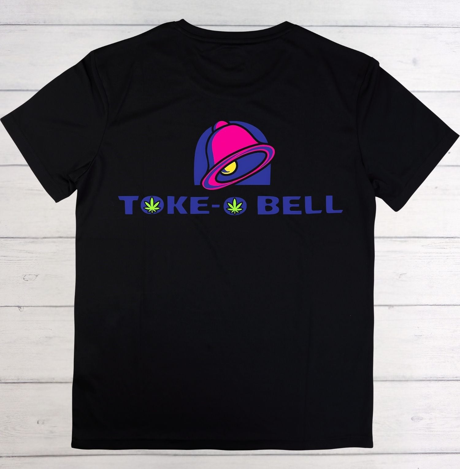 A black short-sleeve t-shirt laid flat on a white wood plank background, featuring a colorful graphic of a bell with a green eye and the text 'TOKE BELL' in blue with green leaf icons.