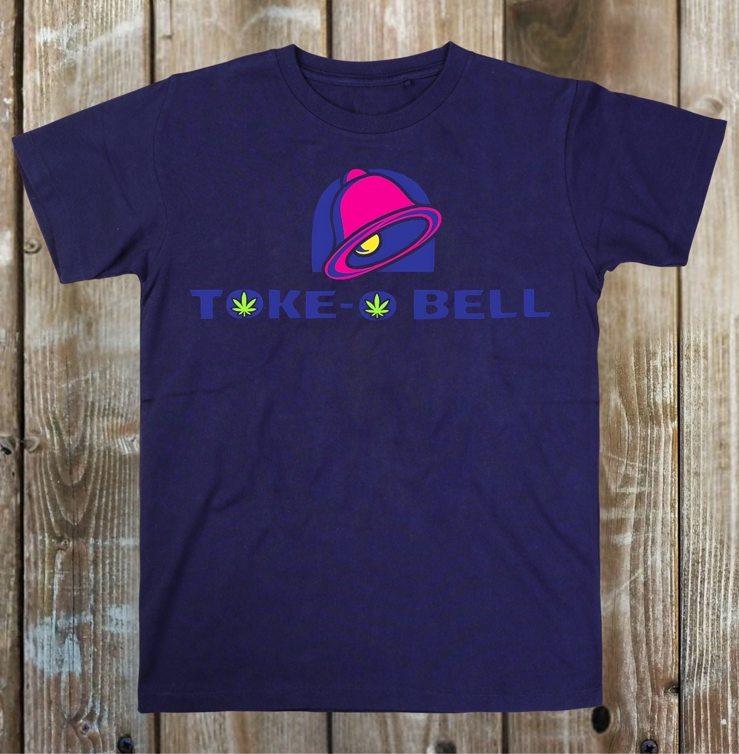 A navy blue short-sleeve t-shirt with a graphic print on the front, laid flat on a rustic wooden plank background. The graphic features a pink and purple taco bell illustration above the text 'TOKE BELL' in purple, with green marijuana leaves replacing the letter 'O'.
