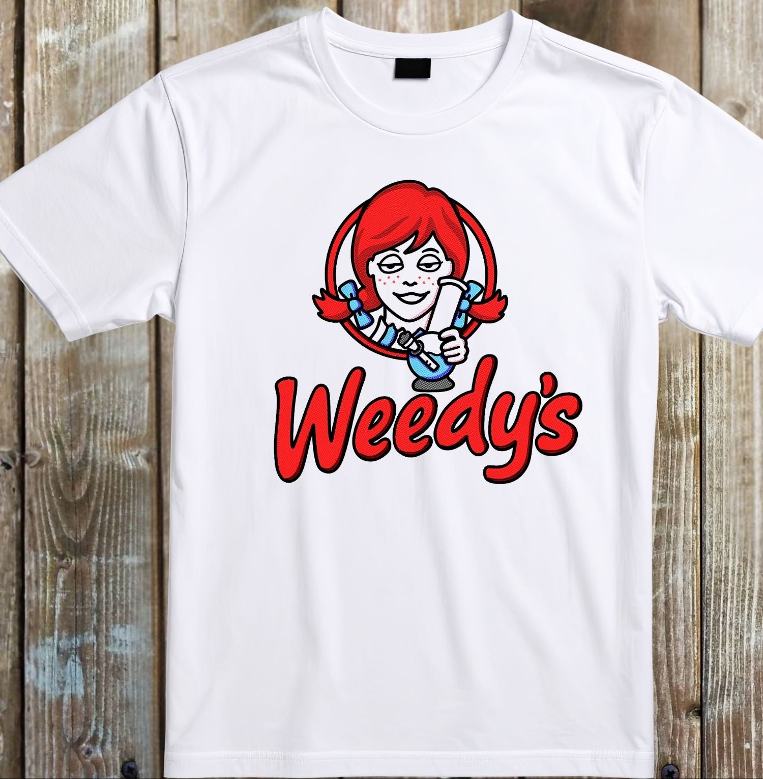 A white short-sleeve crewneck t-shirt featuring a graphic of the fictional character Wendy's red-haired mascot above the red 'Weedy's' text, displayed flat against a rustic wooden plank background.