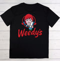 A black short-sleeve t-shirt laid flat on a white wood plank background, featuring a large graphic of the character Wendy from Child's Play with red hair and the word 'Weedy's' in red script.