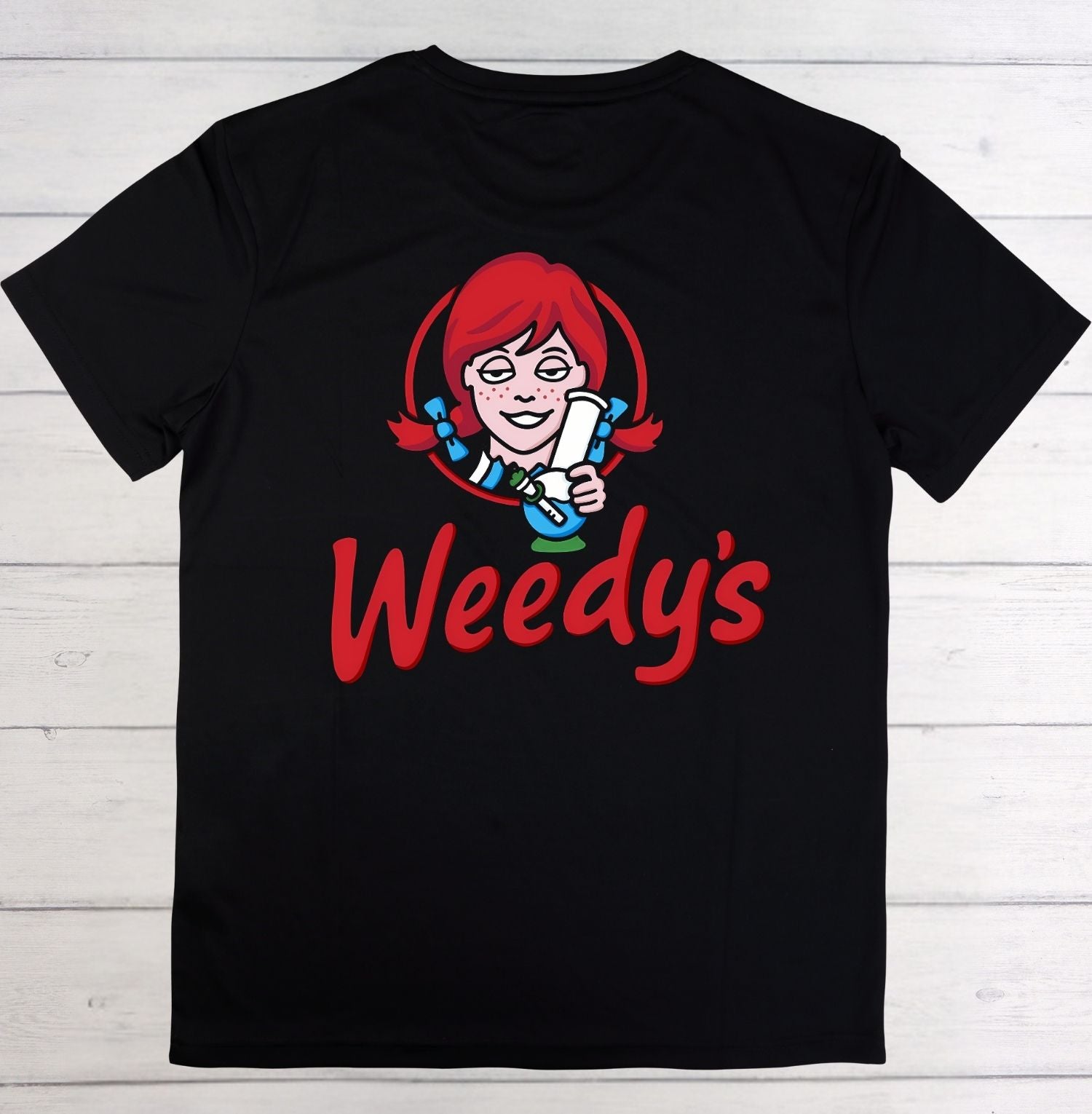 A black short-sleeve t-shirt laid flat on a white wood plank background, featuring a large graphic of the character Wendy from Child's Play with red hair and the word 'Weedy's' in red script.