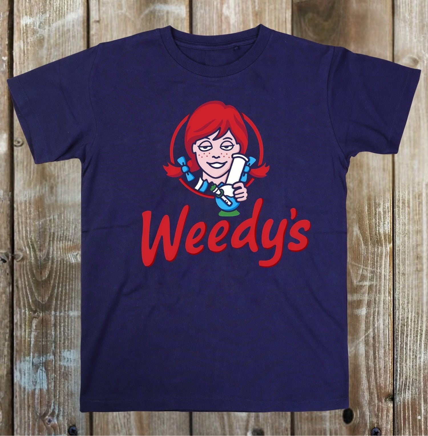A navy blue short-sleeve t-shirt featuring a cartoon graphic of a red-haired woman holding a white pill bottle, with the word 'Weedy's' printed in red below, displayed flat against a rustic wooden plank background.