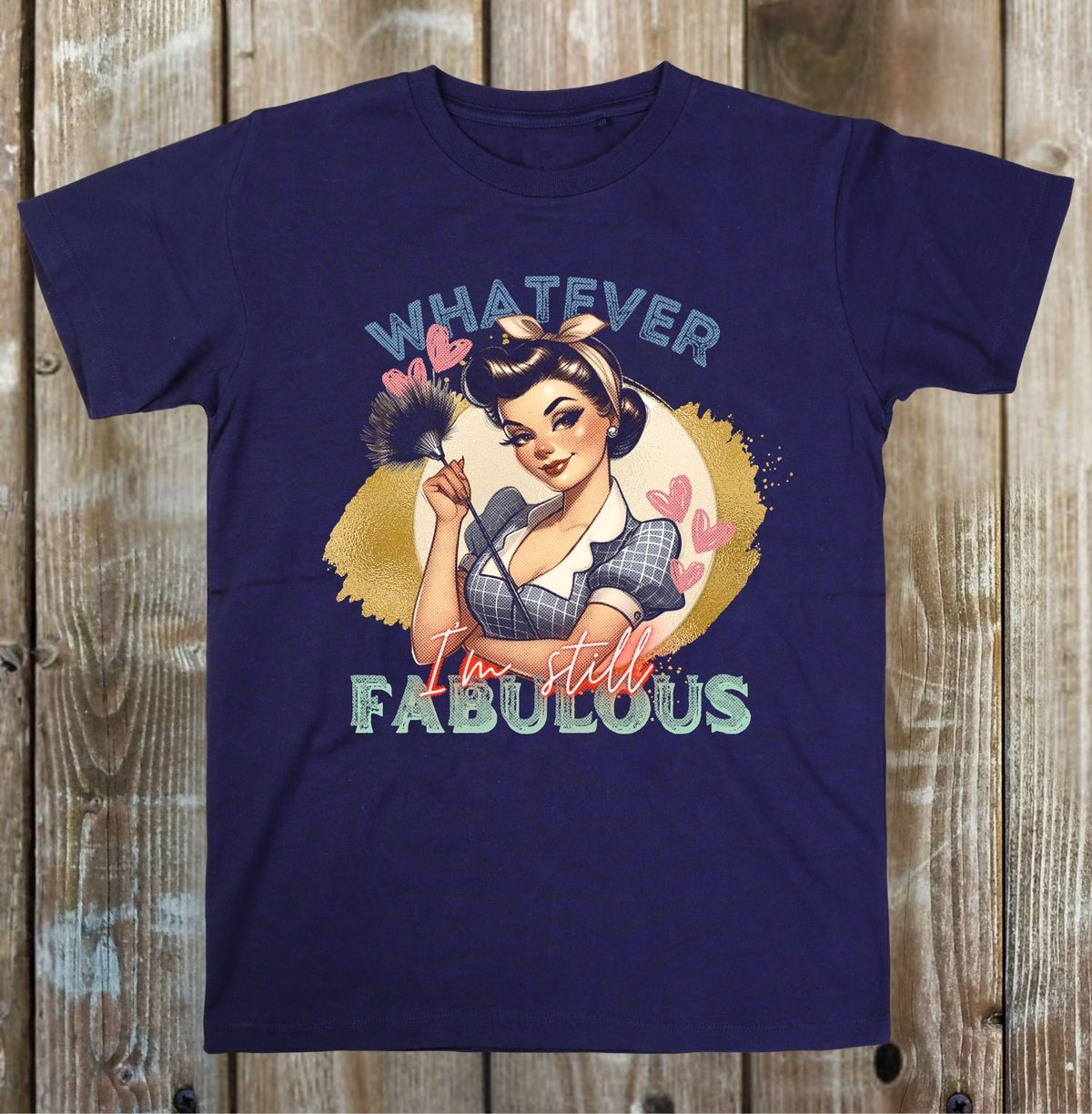 A navy blue short-sleeve t-shirt with a retro pin-up girl graphic and the text 'WHATEVER I'm still FABULOUS' printed on the front, displayed flat against a rustic wooden plank background.