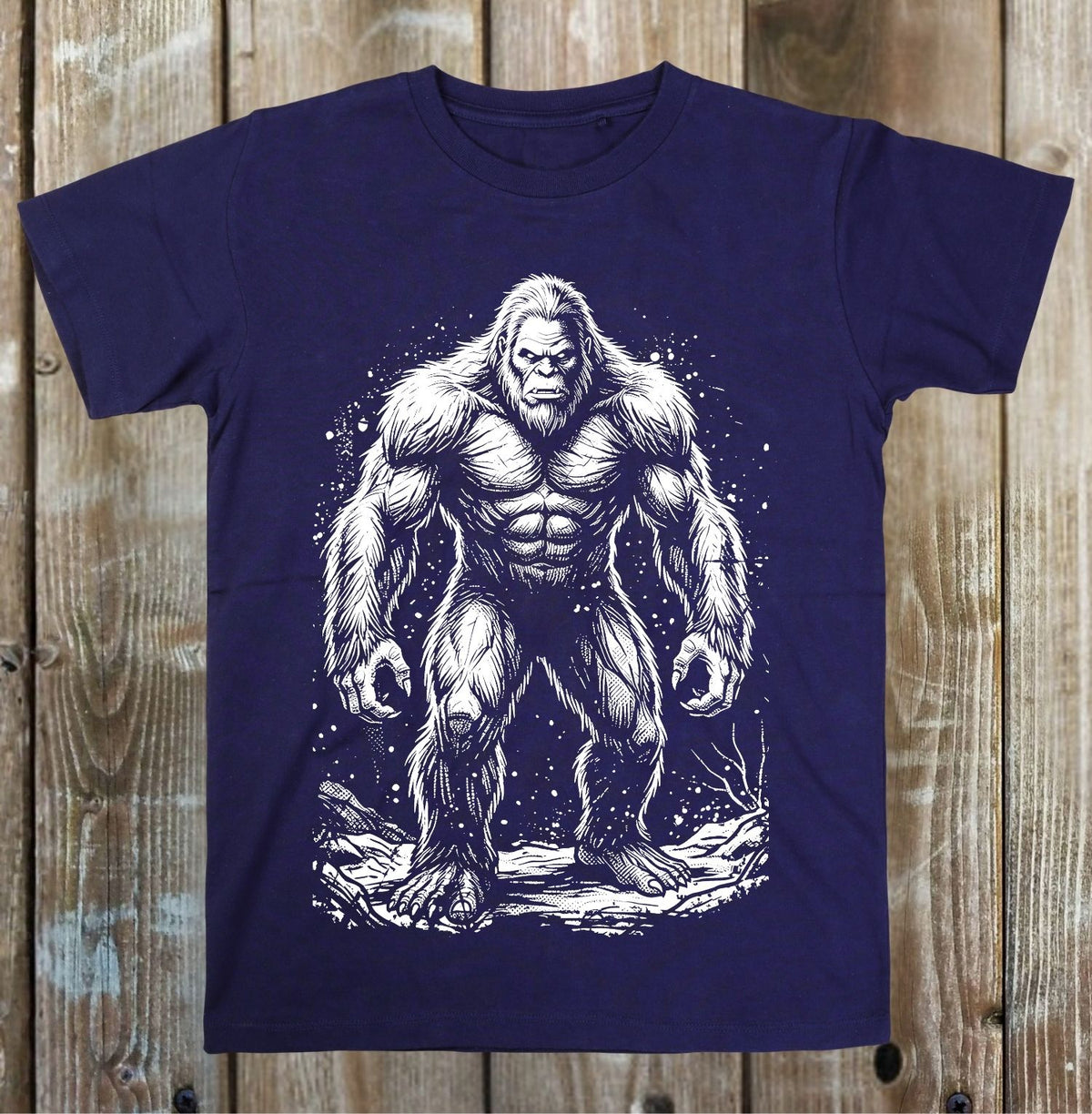 A navy blue short-sleeve t-shirt with a detailed white graphic of a muscular Bigfoot standing on a tree stump, displayed flat against a rustic wooden plank background.